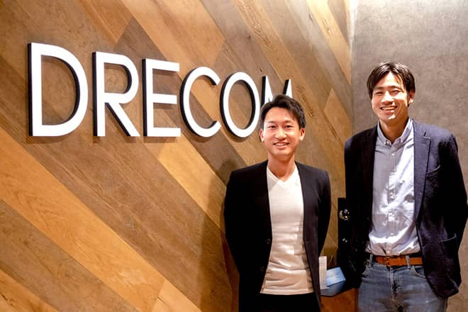 株式会社ドリコムのロゴの前に立っている2人の人